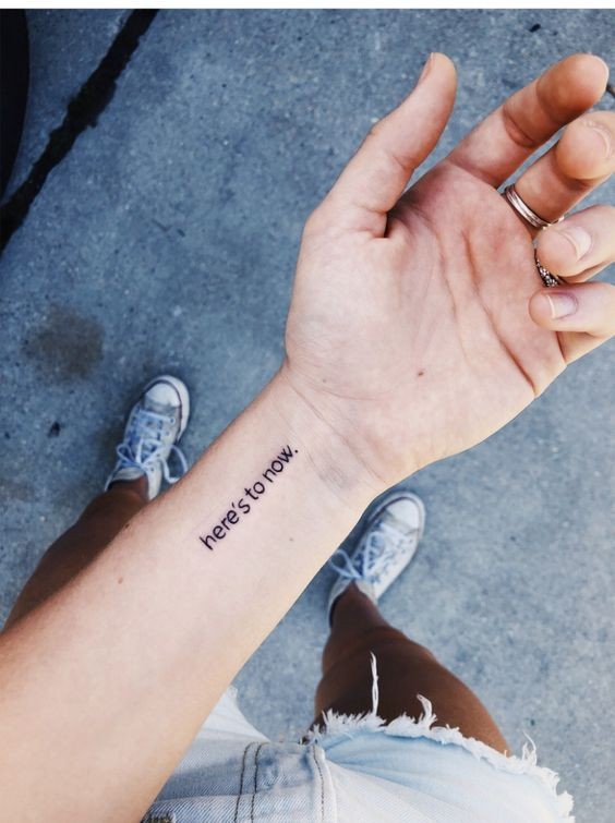 Hình xăm chữ "Here's to now" trên cổ tay